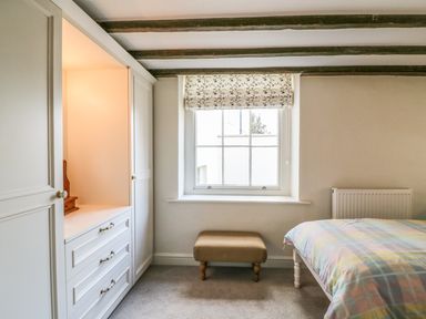 A bedroom with a window a small padded bench a bed with a plaid blanket and built in wardrobes at Tick Tock Cottage in Marnhull