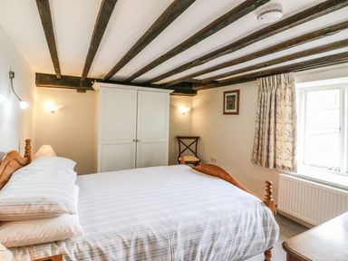 A bedroom with a wooden bed a white wardrobe a wooden chair and a window with curtains at Tick Tock Cottage in Marnhull