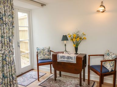 A small seating area with two wooden chairs with floral cushions a small wooden table with a lamp and flowers beside a glass door with floral curtains at Tick Tock Cottage in Marnhull