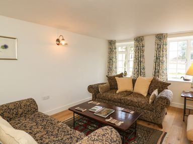 A living room with patterned sofas a wooden coffee table and windows with floral curtains at Tick Tock Cottage in Marnhull
