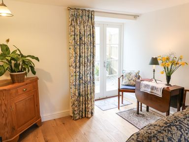A room with wooden cabinet, floral curtains, wooden chair with cushion, small table with lamp and flowers at Tick Tock Cottage in Marnhull