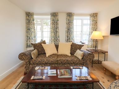A living room with a patterned sofa and cushions a wooden coffee table with books and coasters a side table with a lamp two windows with patterned curtains at Tick Tock Cottage in Marnhull