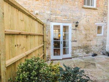 A stone exterior wall with white framed glass double doors next to a wooden fence and plants at Tick Tock Cottage in Marnhull