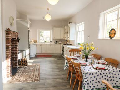 A kitchen with white cabinets a dining table set with dishes and yellow flowers a brick fireplace and windows letting in light at Tick Tock Cottage in Marnhull