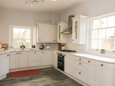 A kitchen with white cabinets two windows and two oval rugs on the floor at Tick Tock Cottage in Marnhull