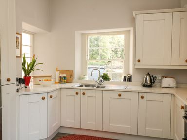 A kitchen with white cabinets a sink under a window a kettle a toaster and a plant on the counter at Tick Tock Cottage in Marnhull