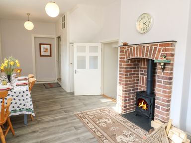 A dining area with a table, wooden chairs, a vase with flowers, and a brick fireplace with a wood-burning stove at Tick Tock Cottage in Marnhull