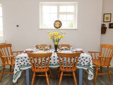 A dining table with six wooden chairs set with bowls glasses and a vase of yellow flowers at Tick Tock Cottage in Marnhull
