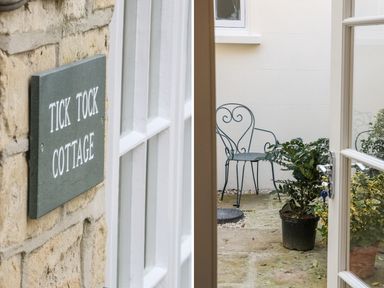 A stone wall with a Tick Tock Cottage sign and an open glass door leading to a small outdoor area with a metal chair and potted plants at Tick Tock Cottage in Marnhull