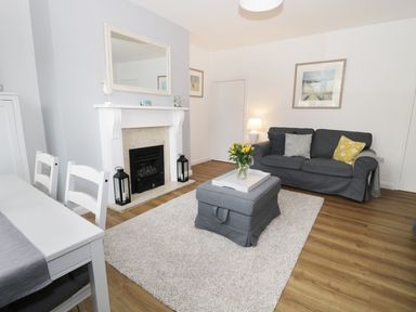 A living room with a gray sofa, a gray ottoman with flowers on it, a white fireplace with two black lanterns, a white dining table with chairs, and framed pictures on the walls at Rose Cottage in Lesbury near Alnmouth