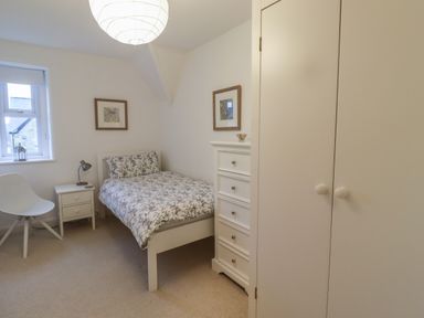 A bedroom with a single bed, a white chair, a small bedside table with a lamp, a chest of drawers, and two framed pictures on the wall at Rose Cottage in Lesbury near Alnmouth