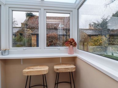 A seating area with two wooden stools and a white windowsill with a vase of flowers and a container of clothespins by a window at Rose Cottage in Lesbury near Alnmouth