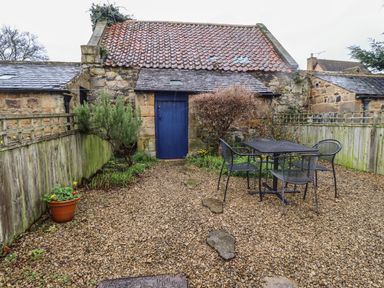 A gravel patio with a metal table and four chairs surrounded by wooden fencing and stone buildings at Rose Cottage in Lesbury near Alnmouth