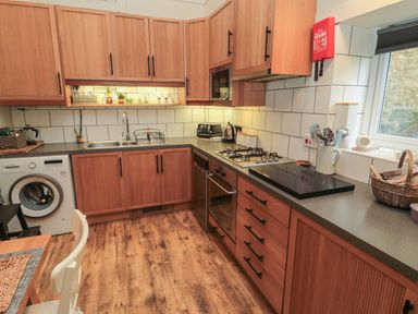 A kitchen with wooden cabinets and drawers a washing machine a gas stove and oven a sink countertop utensils and a window at The Olde Stables in Warkworth