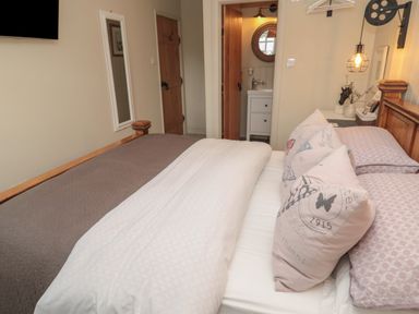 A bedroom with a wooden bed a white and brown bedspread pillows a wall mirror two wooden doors and a small bathroom visible at The Olde Stables in Warkworth