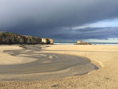 A sandy beach with a small stream flowing to the sea with cliffs and rocks in the background at Chapel Rock View The Dunes in Perranporth