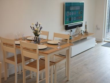 A dining table set with plates glasses and flowers in a room with a wall mounted TV and a wooden cabinet at Chapel Rock View The Dunes in Perranporth