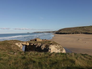 A coastal landscape with a sandy beach cliffs grassy hills and people walking at Chapel Rock View The Dunes in Perranporth