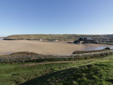 A sandy beach with a grassy hill and fence in the foreground and buildings in the background at Chapel Rock View The Dunes in Perranporth