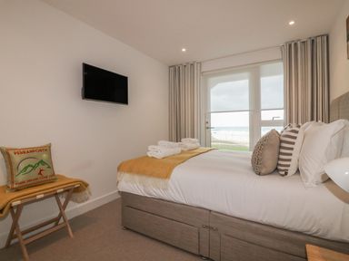 A bedroom with a bed, pillows, blankets, a wall-mounted television, a window with curtains, and a bench with a pillow and blanket at Chapel Rock View, The Dunes in Perranporth