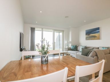 A dining table with a vase of flowers and white chairs in a living room with sofas and a wall painting at Chapel Rock View The Dunes in Perranporth
