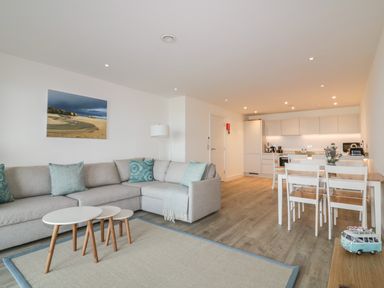 A living room with a gray sectional sofa and nested coffee tables next to a dining table with chairs and an open kitchen at Chapel Rock View The Dunes in Perranporth
