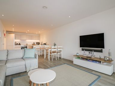 A living room with a grey sofa white round tables a dining table with chairs a wall mounted TV and a kitchen in the background at Chapel Rock View The Dunes in Perranporth