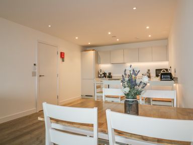 A dining area with a wooden table and white chairs with flowers in a vase and a kitchen with white cabinets and appliances at Chapel Rock View The Dunes in Perranporth