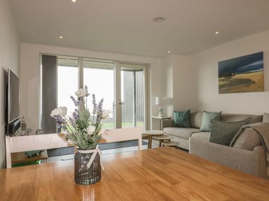 A living room with a wooden table and a vase of flowers next to a gray sofa with cushions at Chapel Rock View The Dunes in Perranporth