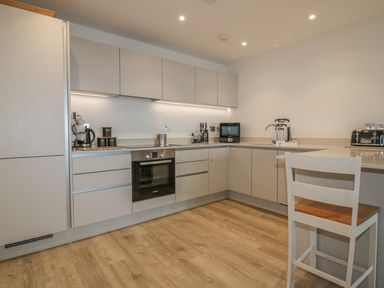 A kitchen with white cabinets an oven a microwave a kettle a toaster and a chair at Chapel Rock View The Dunes in Perranporth