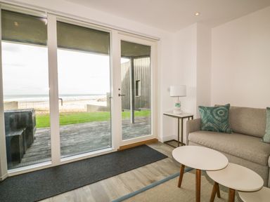 A living room with a sofa a side table with a lamp two nesting coffee tables and glass doors overlooking a garden and beach at Chapel Rock View The Dunes in Perranporth
