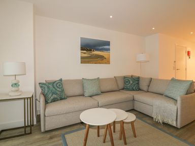 A living room with a beige corner sofa with teal cushions a set of three round nesting tables and two lamps at Chapel Rock View The Dunes in Perranporth