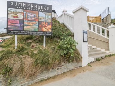 An outdoor sign for Summerhouse bar and restaurant next to a white staircase with a stone wall and sandy ground at Chapel Rock View The Dunes in Perranporth