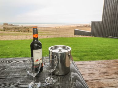 A bottle of merlot wine two empty wine glasses and a metal ice bucket on a wooden table outdoors near a grassy area and beach at Chapel Rock View The Dunes in Perranporth