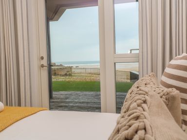 A bedroom with striped curtains and knitted pillows looking out to a beach through glass doors at Chapel Rock View The Dunes in Perranporth