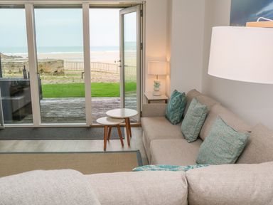 A living room with a beige sofa with blue cushions two nesting tables a lamp and sliding glass doors opening to a beach view at Chapel Rock View The Dunes in Perranporth
