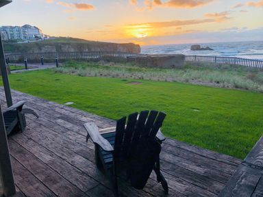 A wooden deck with two chairs facing a grassy lawn and a fenced ocean view at sunset at Chapel Rock View The Dunes in Perranporth
