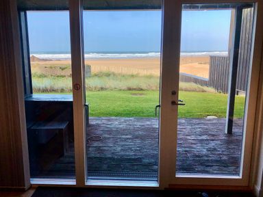 A glass door opening to a wooden deck and grassy area with a sandy beach and ocean in the background at Chapel Rock View The Dunes Perranporth