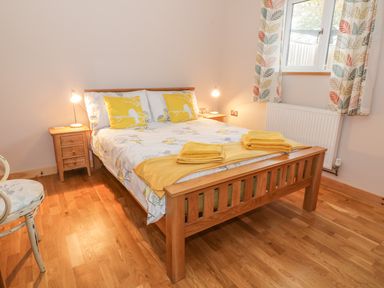 A bedroom with a wooden bed with yellow and white bedding two bedside tables with lamps and a window with leaf patterned curtains at The Stable at Church Farm House Limpenhoe near Reedham