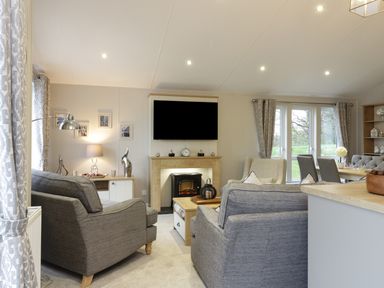 A living room with gray armchairs a TV above a fireplace and a dining area with a table at Woodstock Lodge in Wemyss Bay