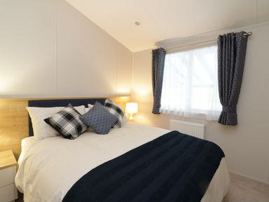 A bedroom with a bed with pillows a bedside table with a lamp and a window with curtains at Woodstock Lodge in Wemyss Bay