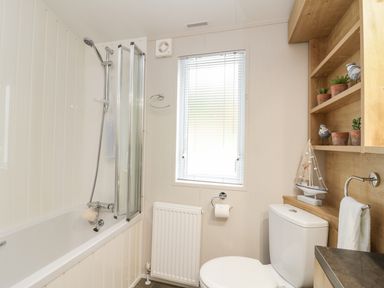 A bathroom with a bathtub and shower beside a window above a radiator and a toilet next to wooden shelves with plants and decorations at Woodstock Lodge in Wemyss Bay
