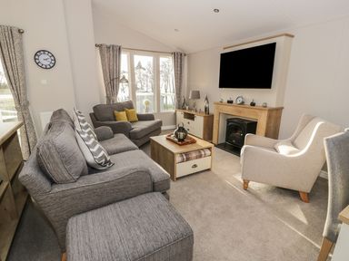 A living room with grey sofas, a wooden coffee table, a fireplace, a wall-mounted television, and large windows at Woodstock Lodge in Wemyss Bay