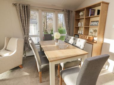A dining room with a wooden table surrounded by grey chairs and a wooden shelving unit by the window at Woodstock Lodge in Wemyss Bay