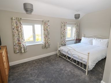 A bedroom with a double bed a wooden chest of drawers two windows with floral curtains and two ceiling lights at 27 Whitegates in Ludham