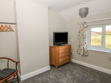 A bedroom with a wooden chair a chest of drawers with a television and a window with floral curtains at 27 Whitegates in Ludham