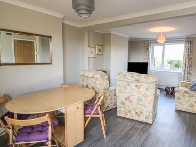 A dining area with a wooden table and chairs next to a living room with floral upholstered chairs and a television at 27 Whitegates in Ludham