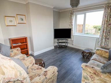 A living room with two floral armchairs a small wooden stool a TV on a glass stand two framed pictures on the wall and floral curtains at 27 Whitegates in Ludham
