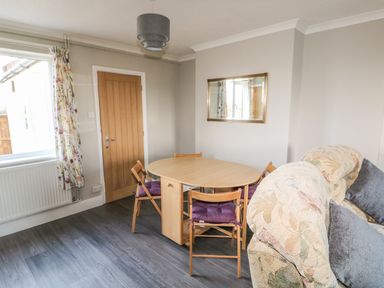 A dining area with a wooden table and four chairs with purple cushions near a floral sofa and a wooden door at 27 Whitegates in Ludham
