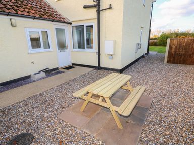 A wooden picnic table on a stone patio outside a yellow house with white windows and a door at 27 Whitegates Ludham